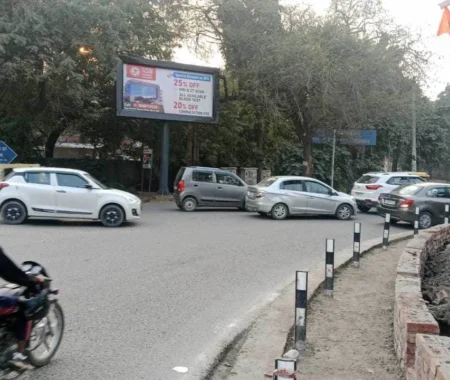 Traffic at a busy roundabout with a roadside billboard advertising 25% off health checkups at SDH Hospital, visible among trees and urban road signage in India.