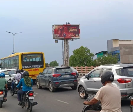 Billboard for Junior Talkies movie content on a congested Indian street with heavy traffic, motorcycles, cars, and a university bus in view.