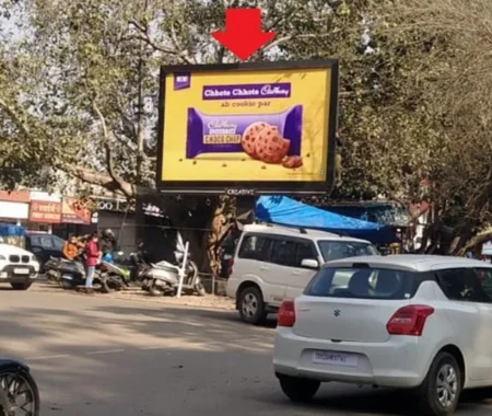 Urban street scene with Cadbury Dairy Milk Chocopie hoarding by ACME Advertising Co., mounted on tree above traffic; features cookie and chocolate bar visual, visible cars, motorcycles, and pedestrians